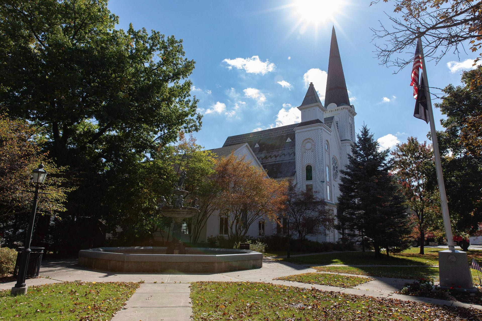 The United Methodist Church in Dryden, NY with its spire reaching upward toward the sun, also in the frame, on the right side of the photo. On the left, there is a tree, and a sidewalk that leads toward the photographer and foreground.