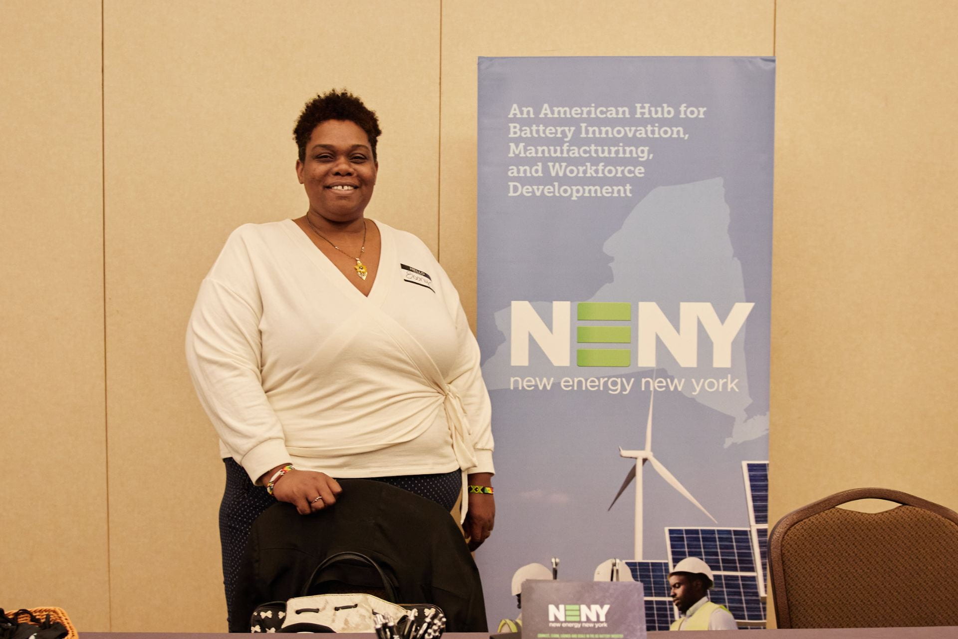 A woman in a white top with a poster that says NENY stands and smiles at the camera from behind a table.