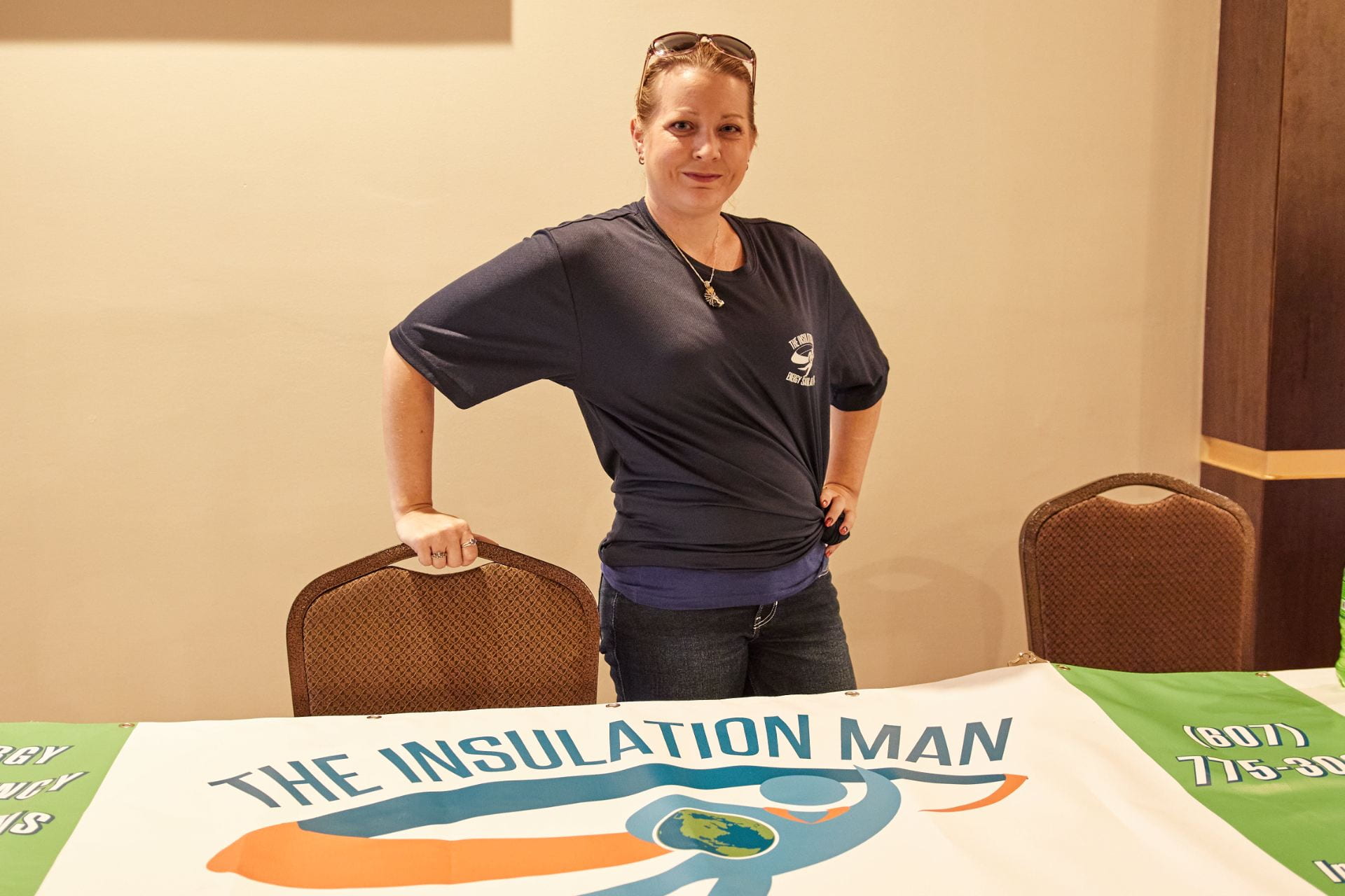 A woman in a blue top with sunglasses on her head smiles at the camera, with a banner on the table below her that reads "The Insulation Man"