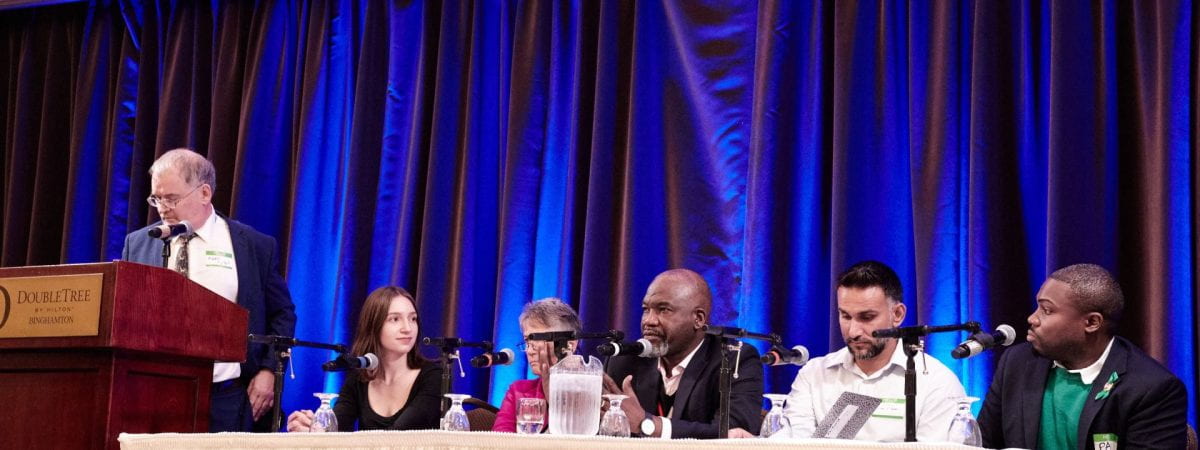 Five people on a panel sit at a table with a white tablecloth, as a man in the middles speaks into the microphone in front of him. The moderator, to the left of the image, stands at a podium and watches and listens.