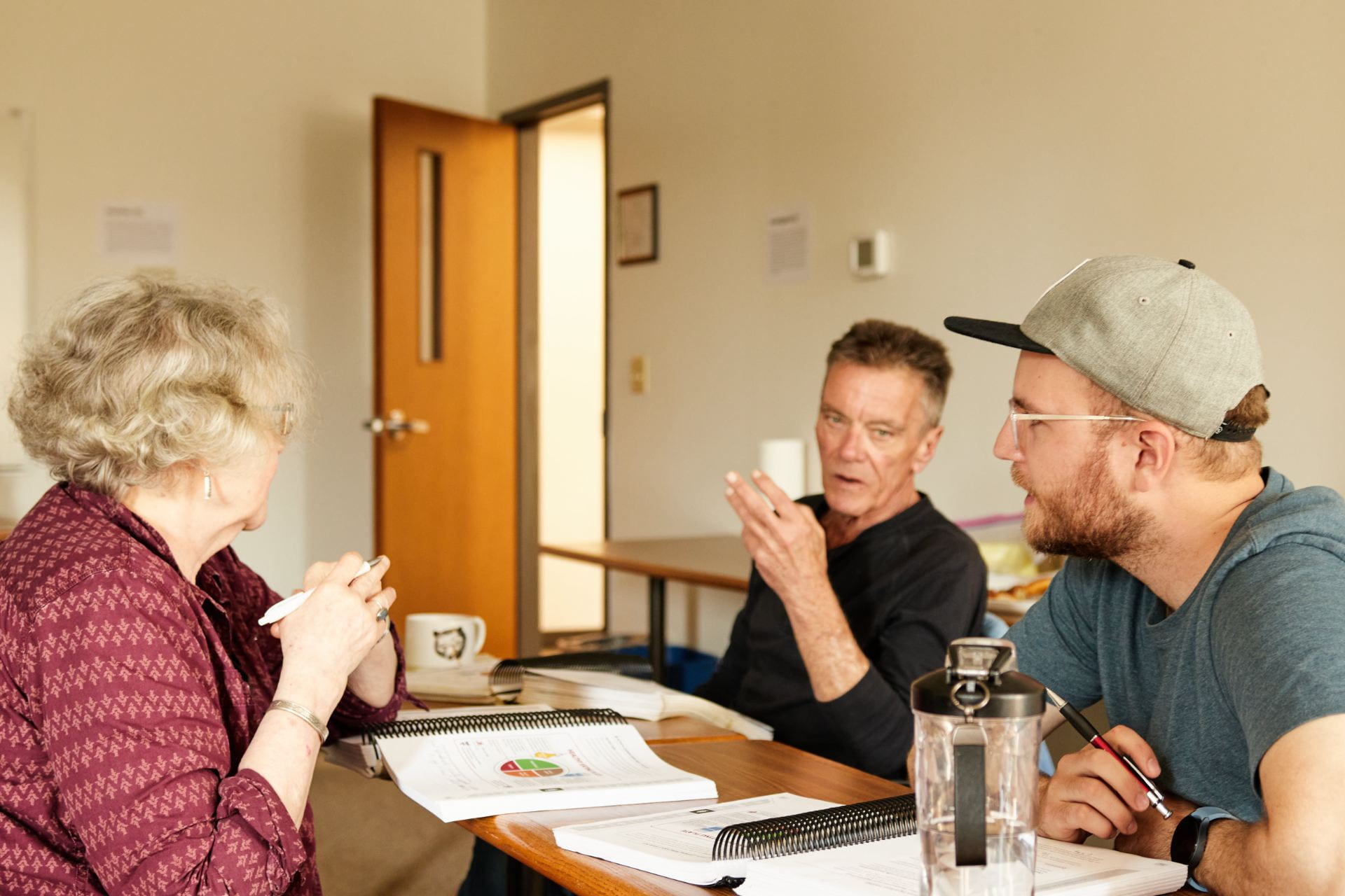 Three students sit in a well-lit classroom, discussing during a group activity. On the left is one student, a woman with curly grey hair, seated across from are two men, one older in a black shirt, speaking with his hand by his face, and another with glasses and a cap, listening.