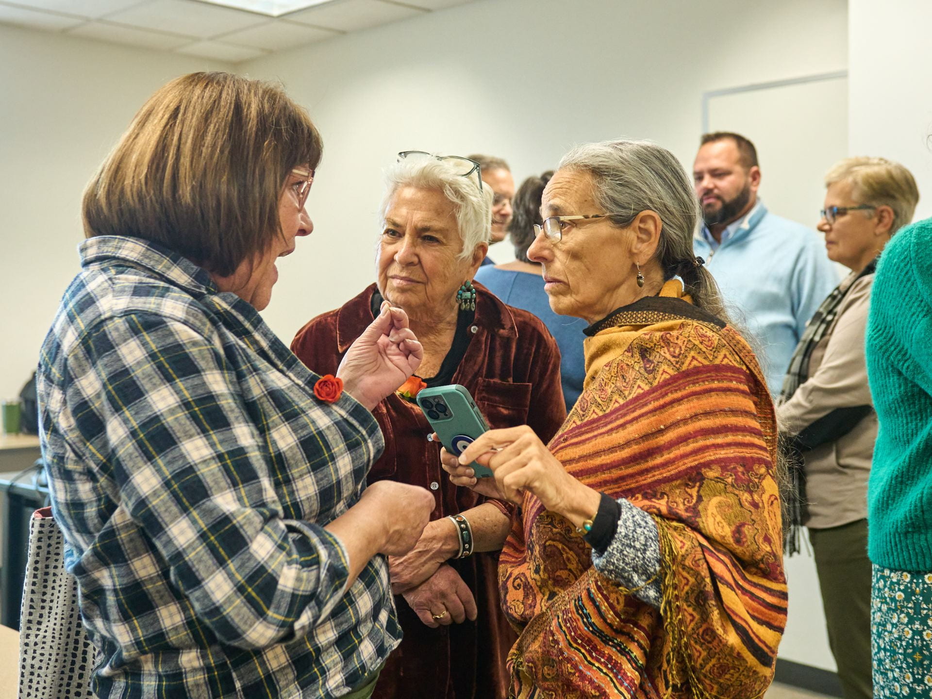 Three women speak in a tight circle inside a well-lit classroom, the one on the left in a grey flannel shirt speaking with her hands as the other two look and listen carefully. A group, out of focus, stands behind in their own circle, having their own conversation
