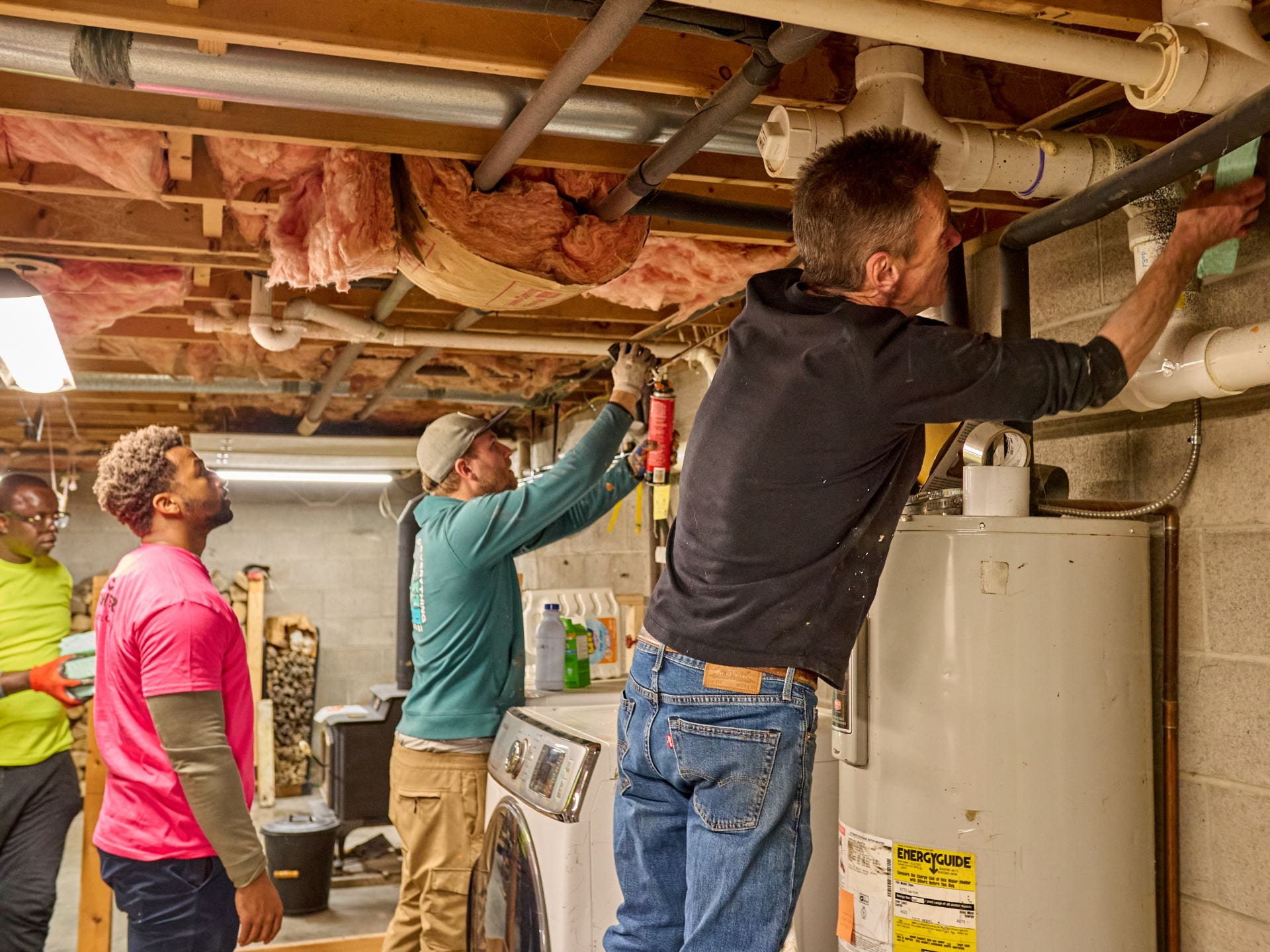 Four men stand on the left side of the frame, looking and working with their hands on the rim joist in the top right of the frame in a well-list basement with insulation overhead